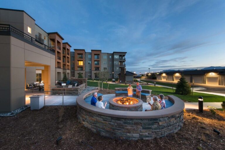 Seniors relaxing around an outdoor fire pit at a modern independent living retirement community at dusk.