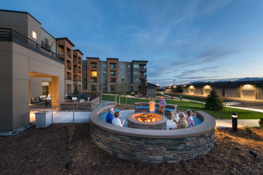 Seniors relaxing around an outdoor fire pit at a modern independent living retirement community at dusk.