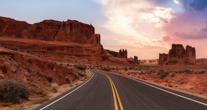 Paved road winding between towering red sandstone formations beneath a pink and purple sunset sky